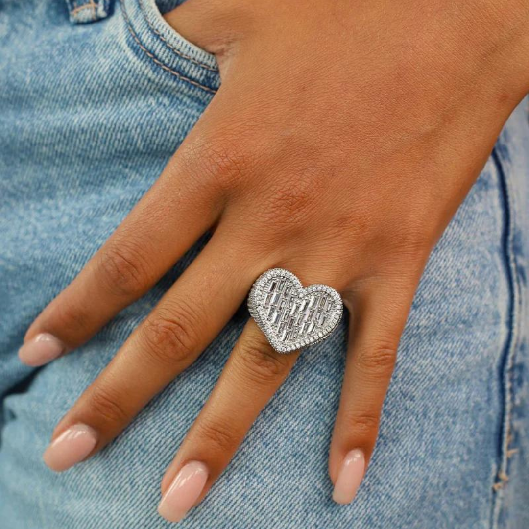 Model's hand showcasing a white gold baguette heart ring, with the heart-shaped design prominently displayed on the finger, set against a stylish, urban background.